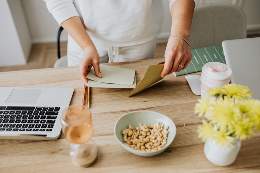 A person arranging an envelope beside a laptop on a sunlit wooden desk